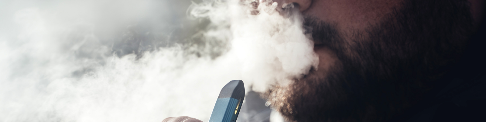 A bearded man exhales vapour from a vape pen device.